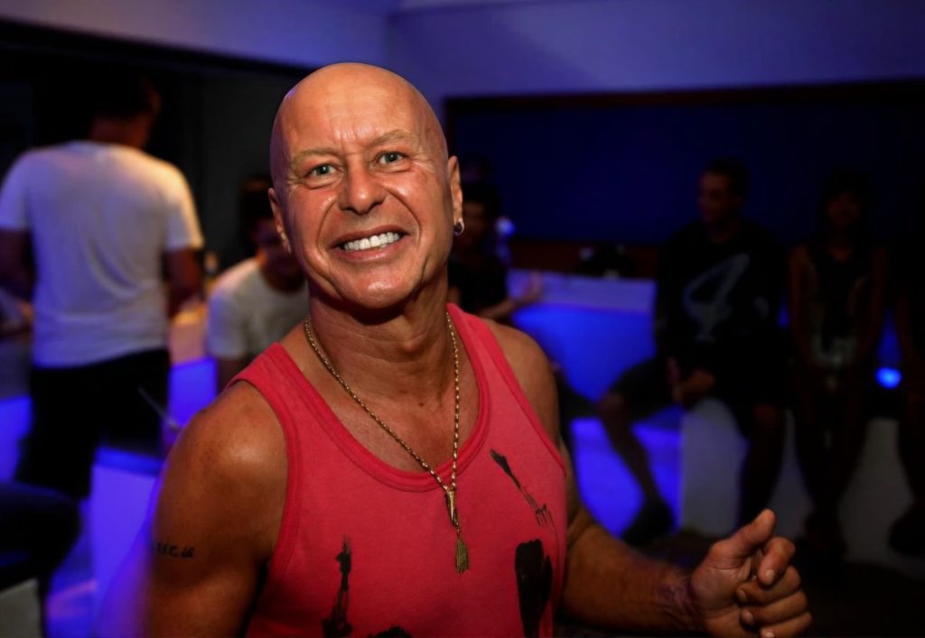 British DJ and producer Graham Gold smiling in a nightclub setting, wearing a pink sleeveless top with blue ambient lighting and people gathered in the background.