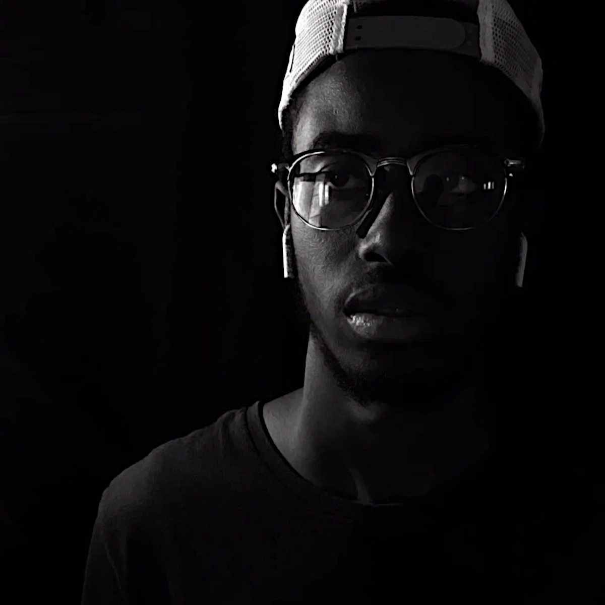 Mozambican DJ and producer Navaa wearing a baseball cap backwards and glasses with small in ear headphones in an upper body shot taken on his right side, black and white image.