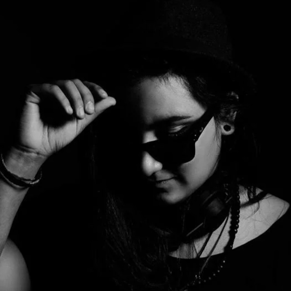 Peruvian DJ Yamile Kouri standing in a hat with her hand on the brim wearing sunglasses, in darkened room, black and white image.