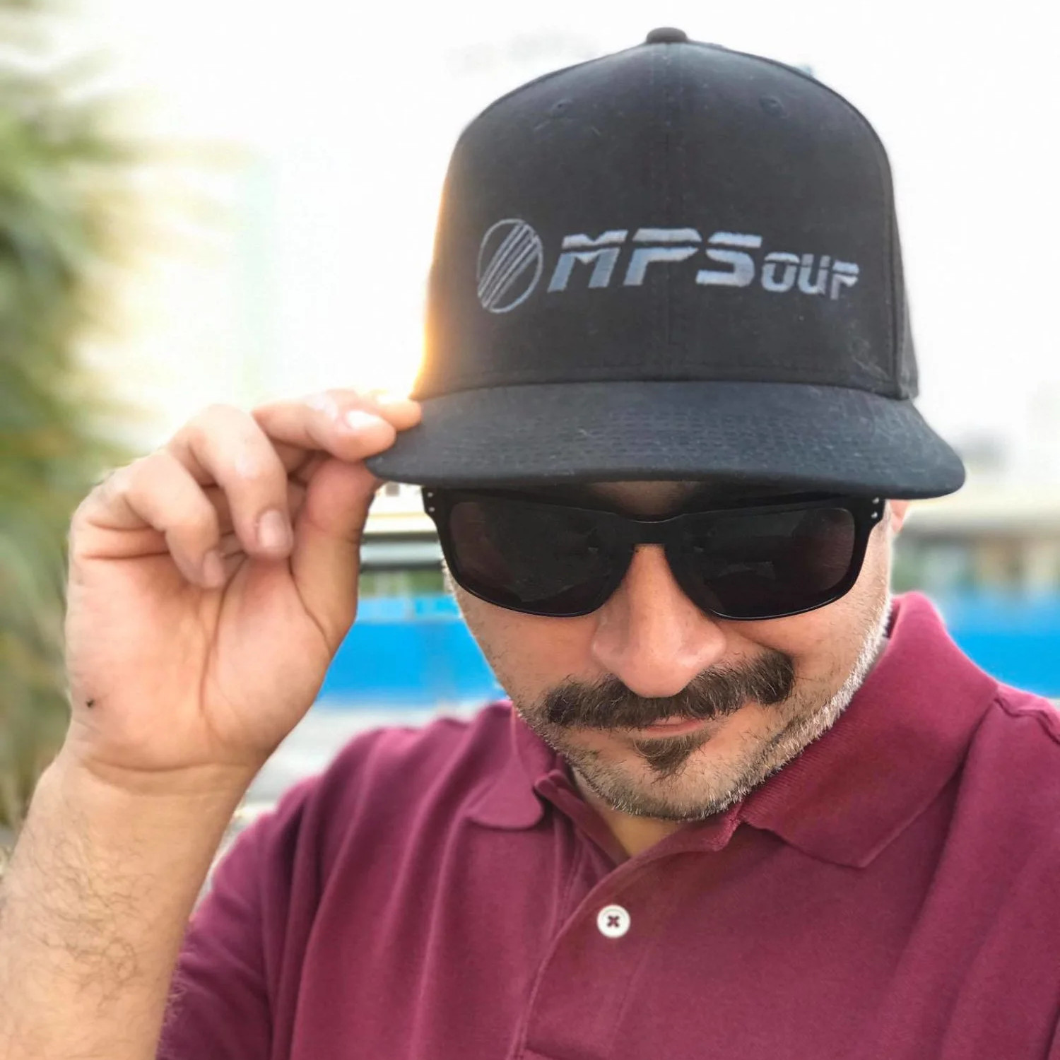 American dJ MPSoup lookimng at camera weeraing sunglasses and a cap with his logo with his right hand on the peak, colour image.