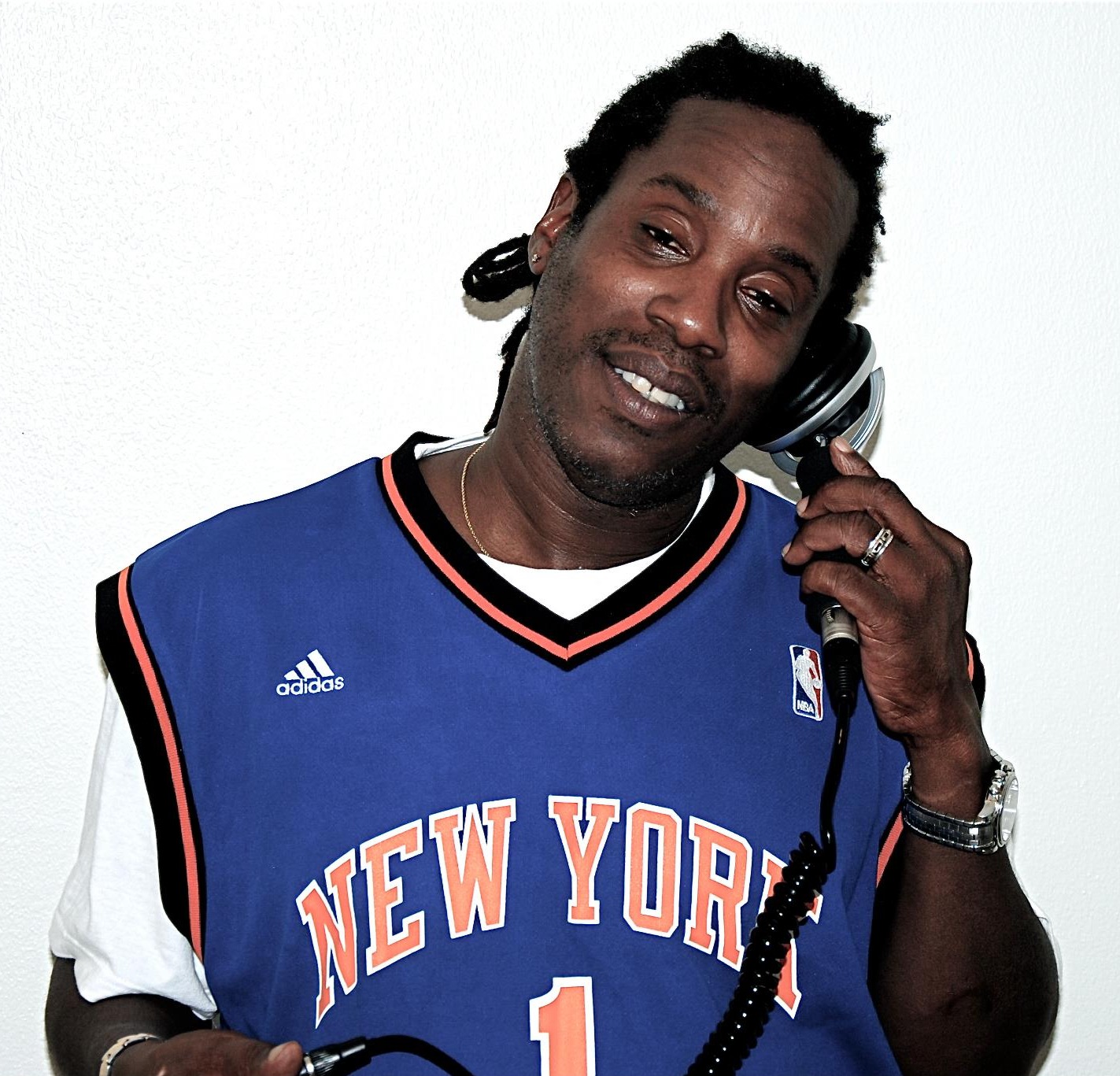British DJ and producer Bert Bevans standing holding a headphone to his left ear and wearing a New York Knicks basketball shirt, colour image.