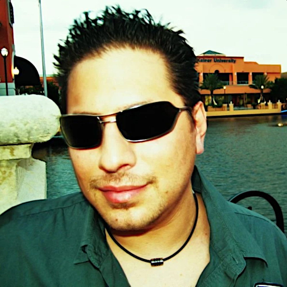 American DJ, producer and record label owner Eddie Lopez posing in a headshot wearing sunglasses with river behind him, colour image