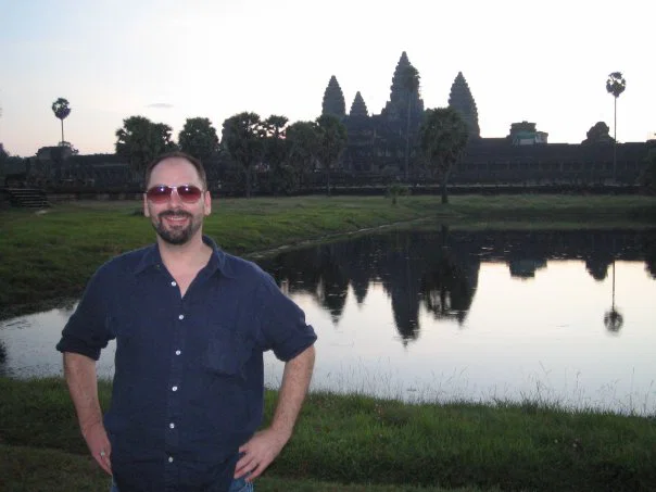 Senior journalist Peter Cornish pictured near Angkor Wat in Cambodia, standing beside a reflective pond with the temple silhouette in the background.
