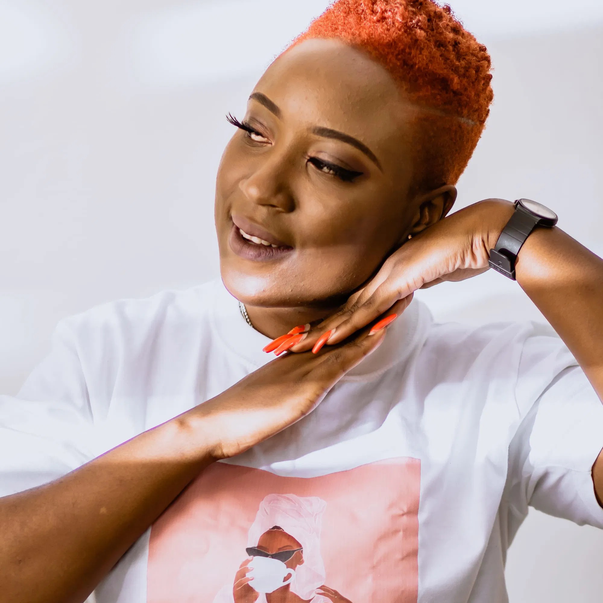 Zambian DJ Ms Selfie with her hands placed together on her left cheek and looking to her right with short dyed hair and a printed white t-shirt, colour image.