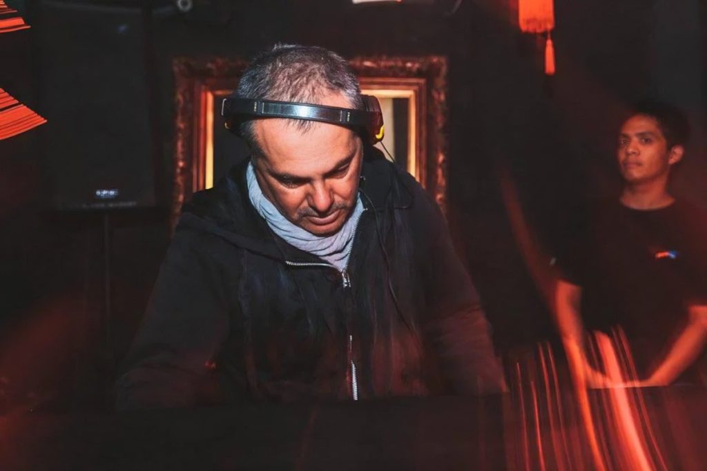 Marco Mei, Italian DJ and producer, focused over the decks wearing headphones in a dark red-lit club setting.