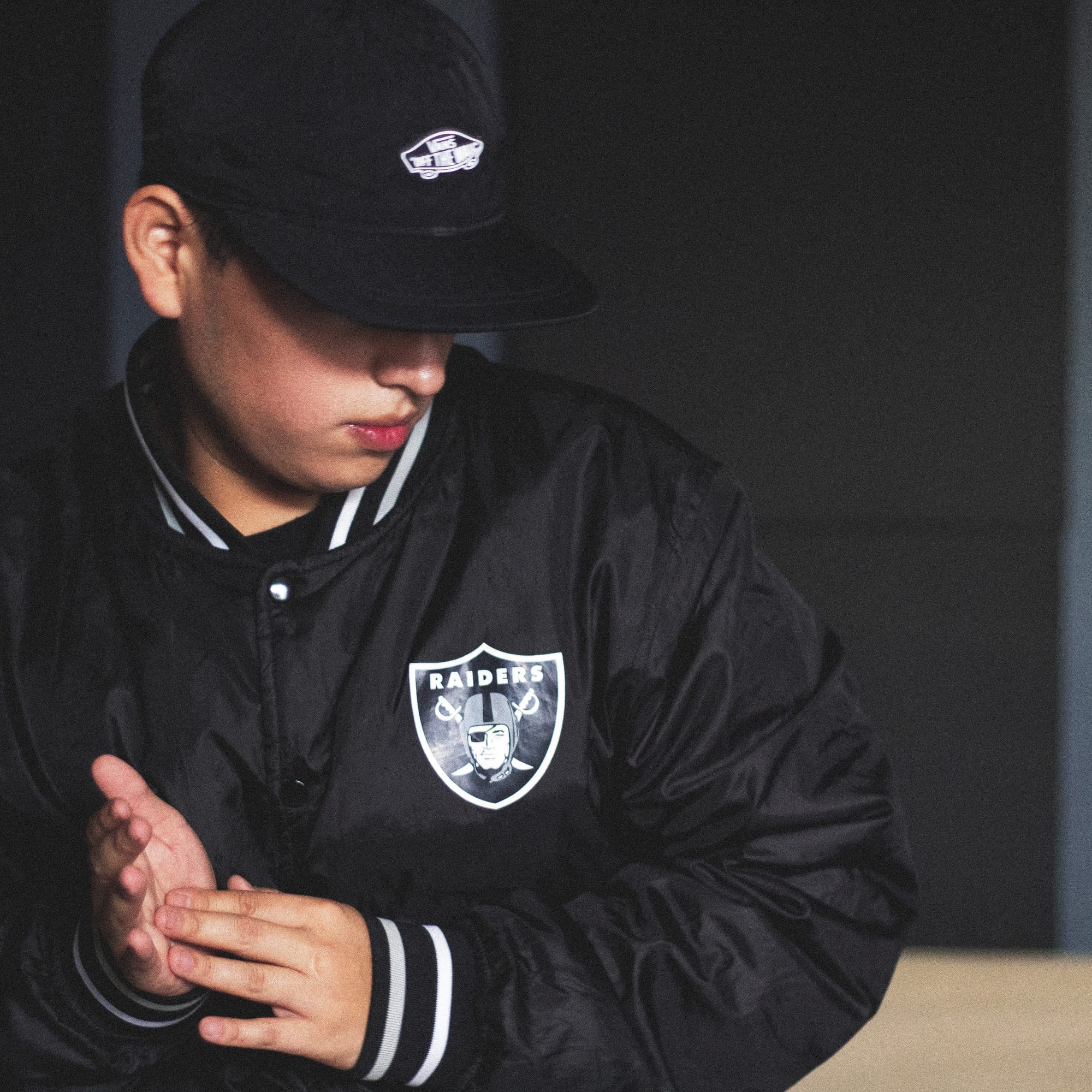 Guest DJ Jizz wearing a black Las Vegas Raiders jacket and black cap with a white car design, looking down and clasping hands against a dark background.