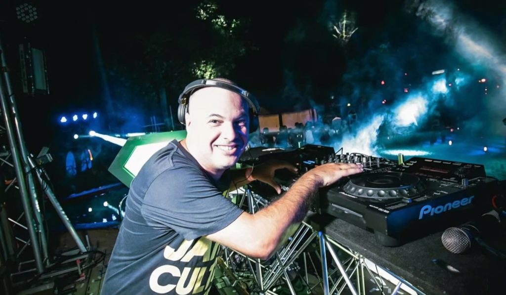 Marco Mei, Italian DJ and producer, smiling at the camera behind Pioneer CDJs at an outdoor festival stage with lights and smoke effects behind him.