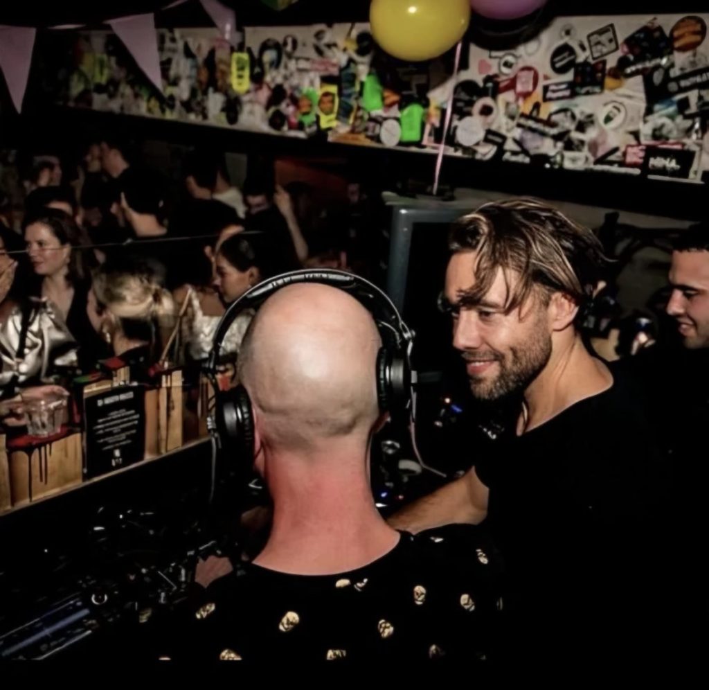 Dutch DJs Reinoud van Toledo and Jack Delhi talk together behind a DJ booth during a busy club night, representing their shared connection through Amsterdam’s underground electronic music scene.
