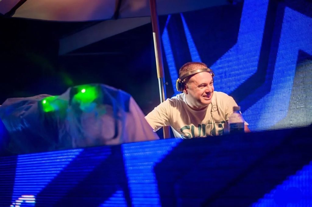 Marco Mei, Italian DJ and producer, grinning behind the decks at a large venue with green lasers and blue stage lighting.