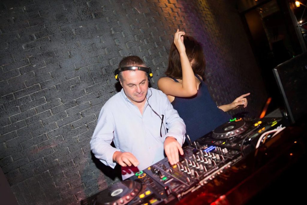 Marco Mei, Italian DJ and producer, focused at the Pioneer CDJs while a dancer moves behind him against an exposed brick wall.