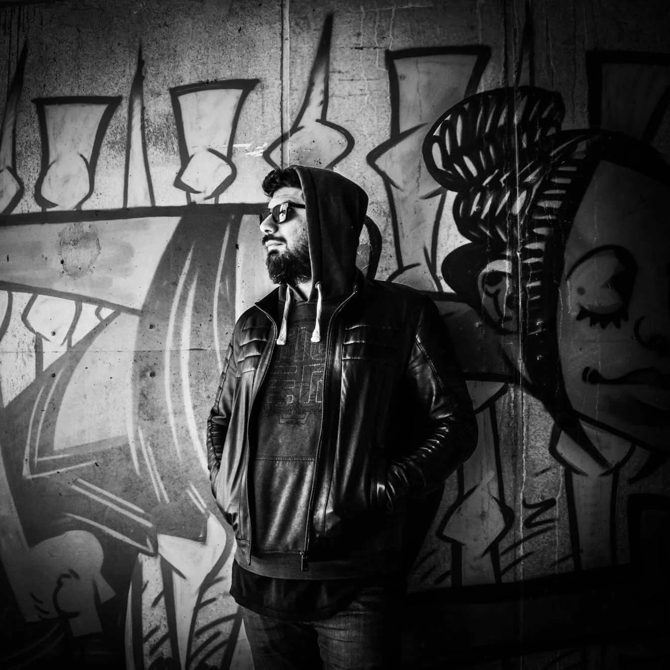 Black-and-white portrait of electronic music artist Concat standing in front of a graffiti-covered wall, wearing sunglasses, a hooded top and a leather jacket while looking to the left.