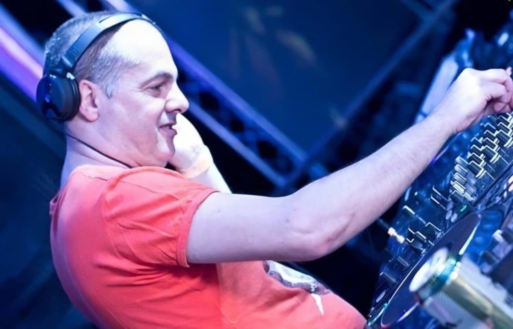 Marco Mei, Italian DJ and producer, smiling at the mixer in a red t-shirt under blue club lighting, shot from the side.