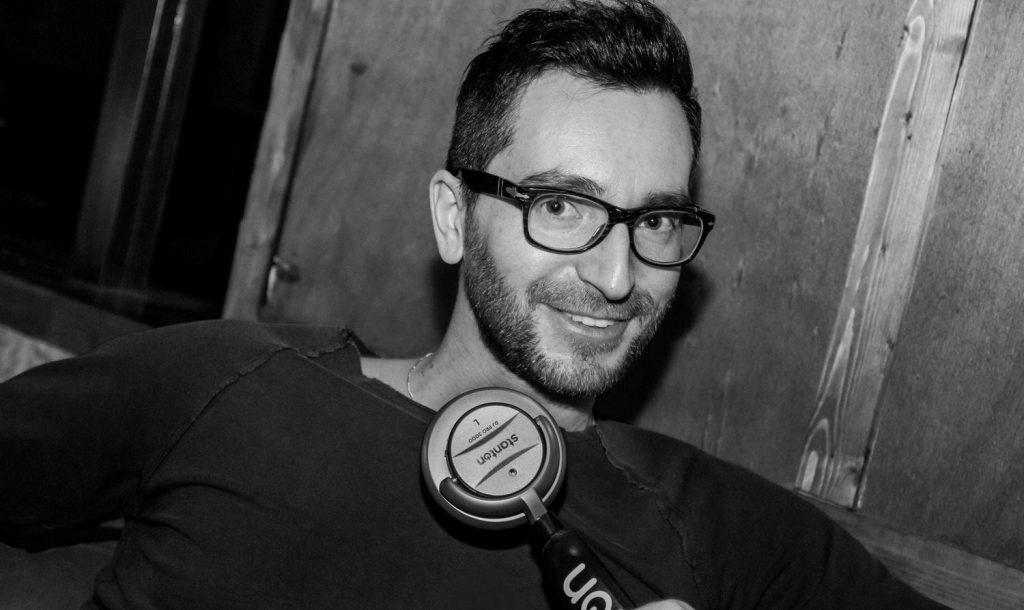 Italian DJ and producer Inve sitting in chair with monophone on his chest and smiling, black and white image.