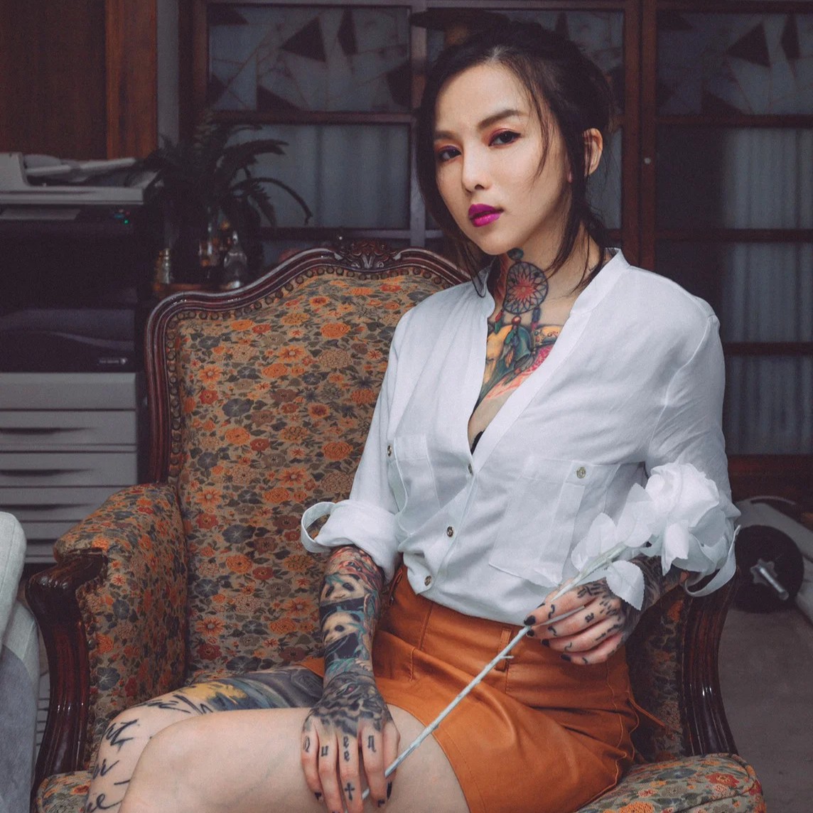 DJ HER, Taiwanese techno DJ and producer, seated in a floral armchair wearing a white shirt, with extensive tattoos visible on her arms, hands and neck.