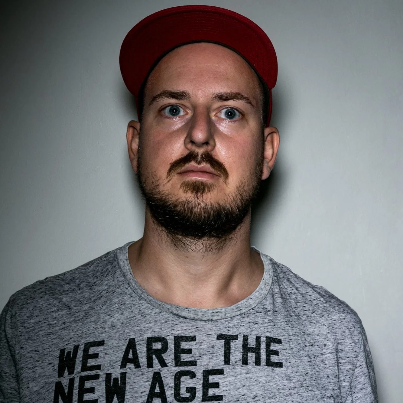 Portrait of Düsseldorf-based Techno DJ and producer Rob van Hell wearing a red cap and grey “We Are the New Age” T-shirt against a plain wall, reflecting his raw and functional approach to underground club music.