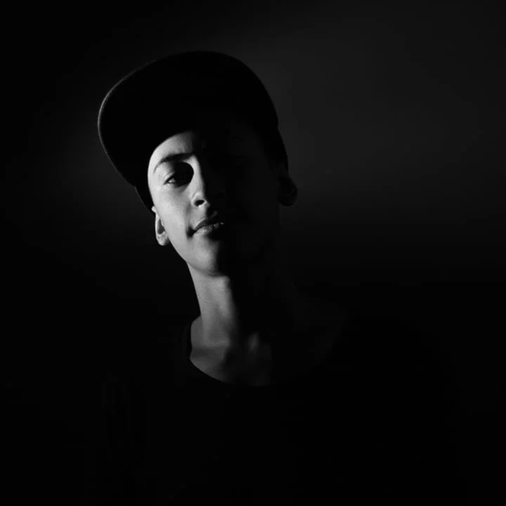 Lucas Romagnoli in a black and white portrait, wearing a cap and gazing upward against a near-black background.