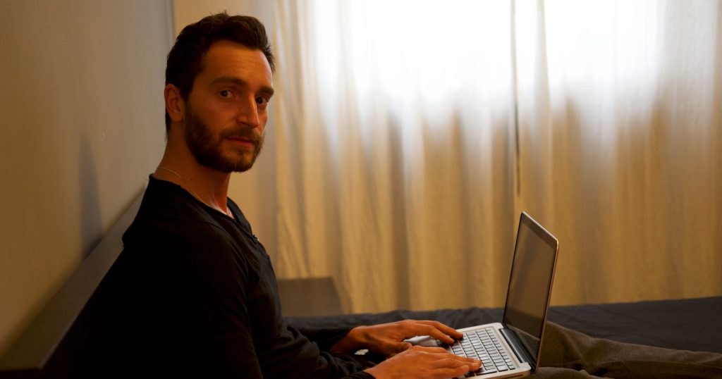 Italian DJ and producer Inve sitting on sofa with legs up working on apple laptop, colour image.