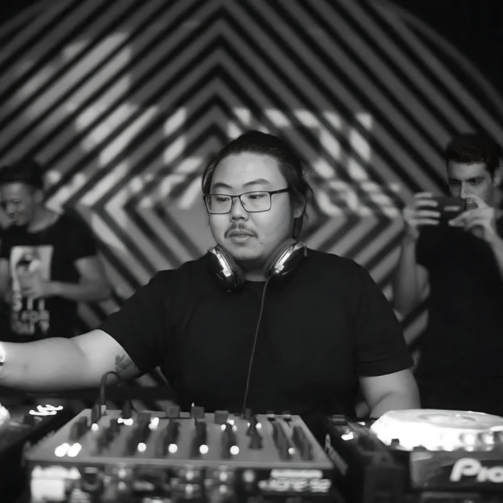 Yudi Watanabe, progressive house DJ and producer from São Paulo, performing at the decks in a black and white shot with a geometric patterned backdrop.