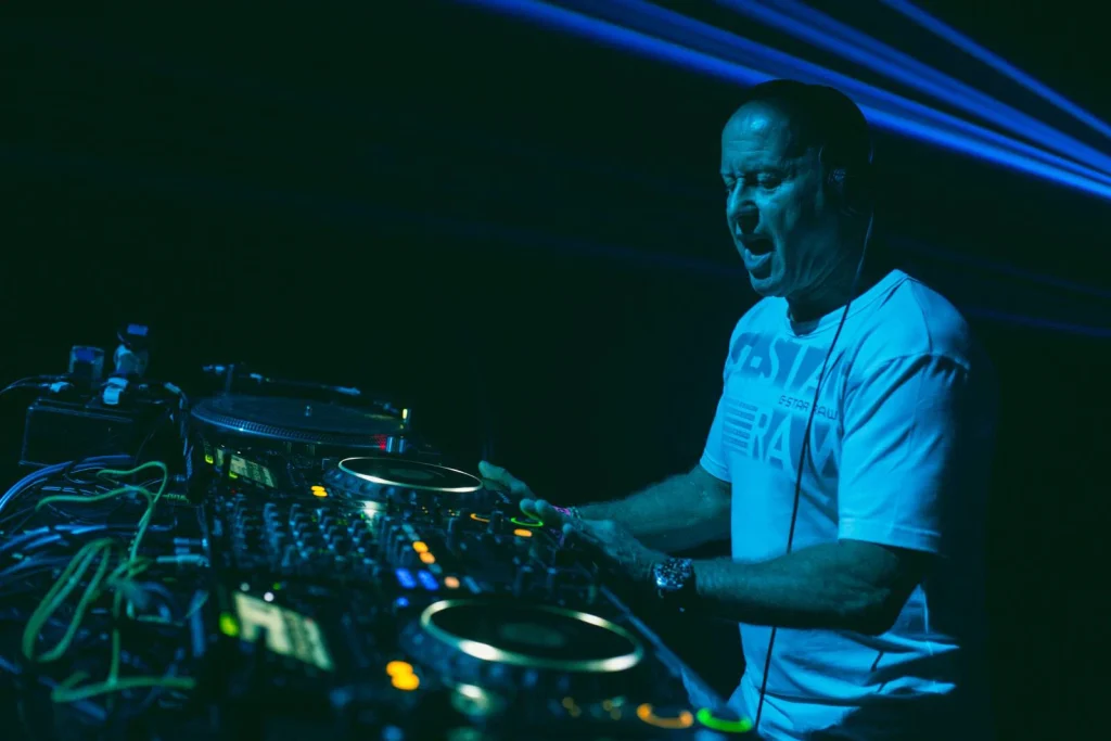 British DJ and producer Graham Gold performing live behind the decks under blue lighting, surrounded by club equipment and laser effects during a nightclub set.