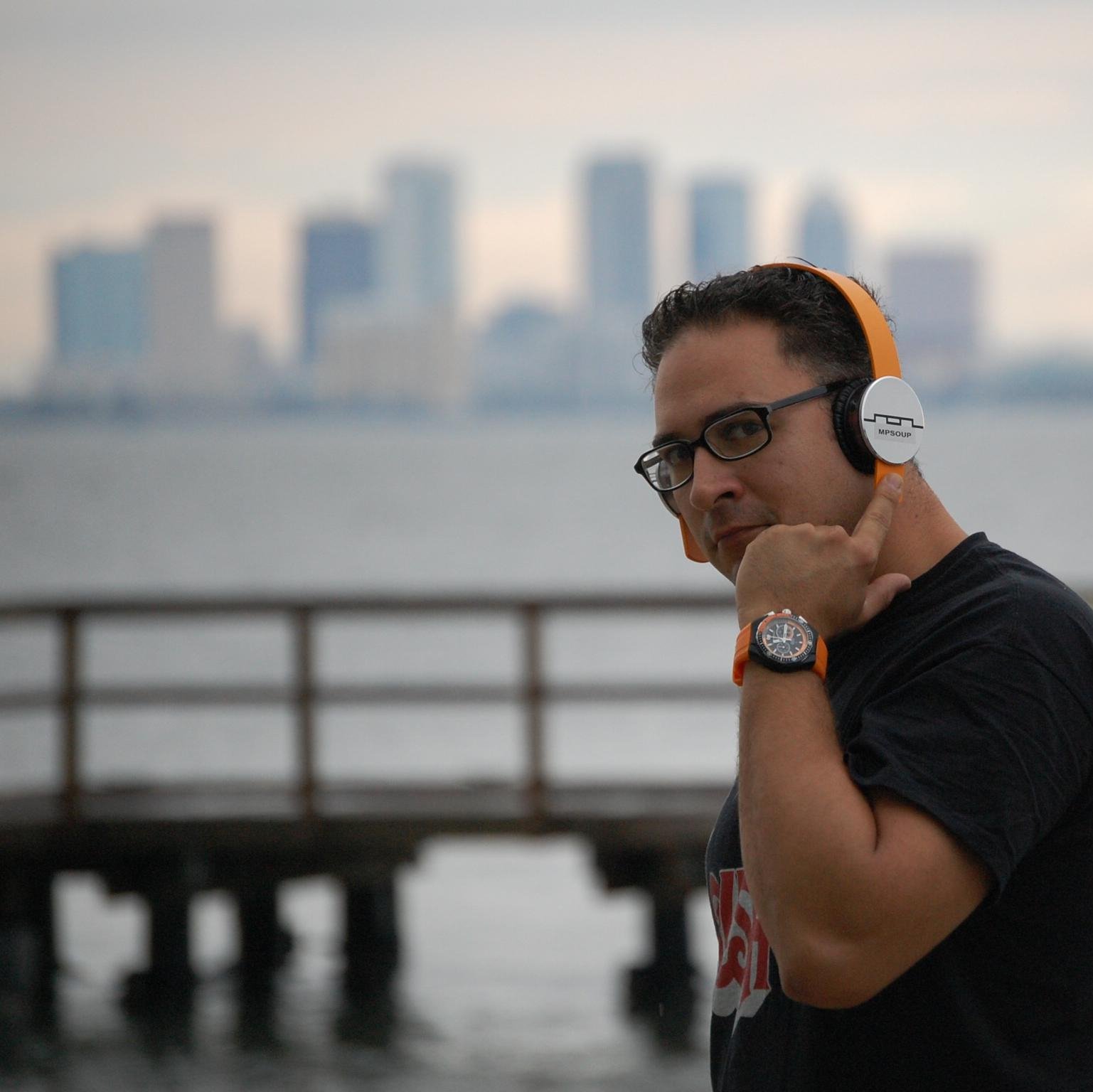 American DJ MPSoup standing side on looking at camera with left hand and finger on headphones on his ears, colour image.