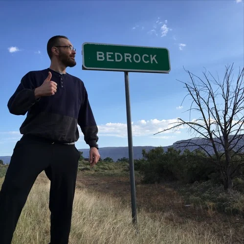 American DJ and producer Andy Pitcher standing in front and looking at a Bedrock roadsign with his thumb up, colour image.