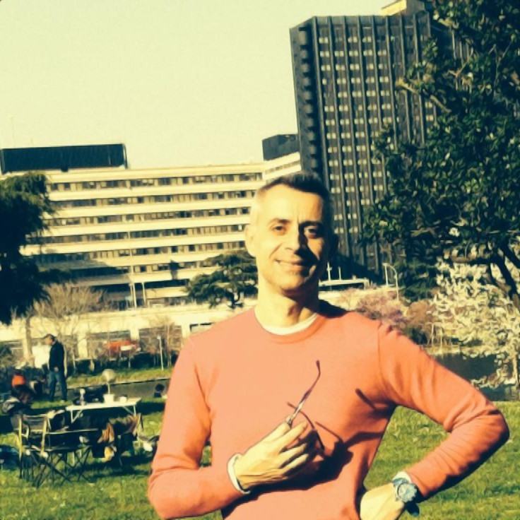 Italian DJ and producer Pier Giorgio Marini standing in park left hand on hip and right hand holding sunglasses, colour image.