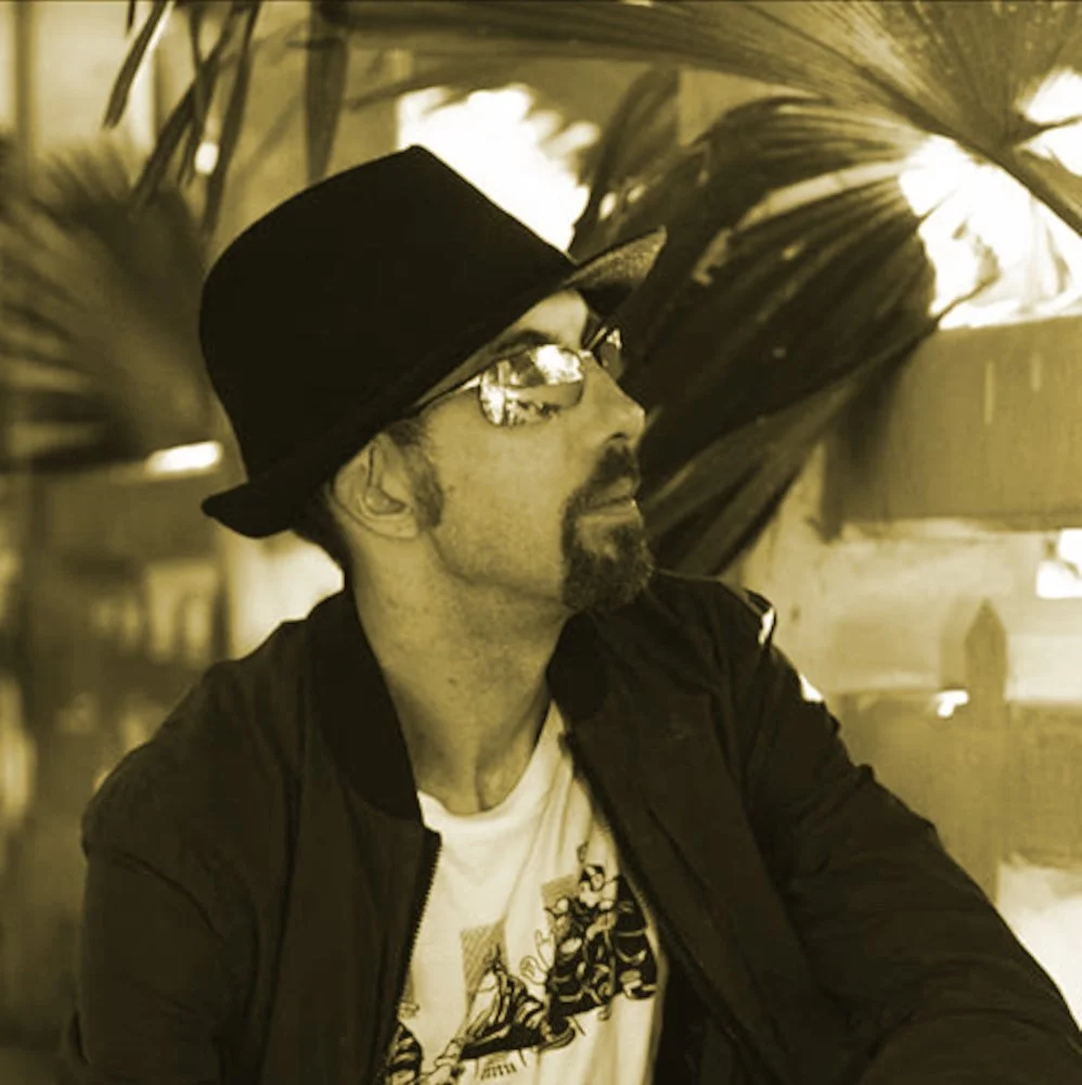 New Zealand DJ Snuck wearing a Hat and Sunglasses looking up to his left whilst sitting, black and white image tinted beige.
