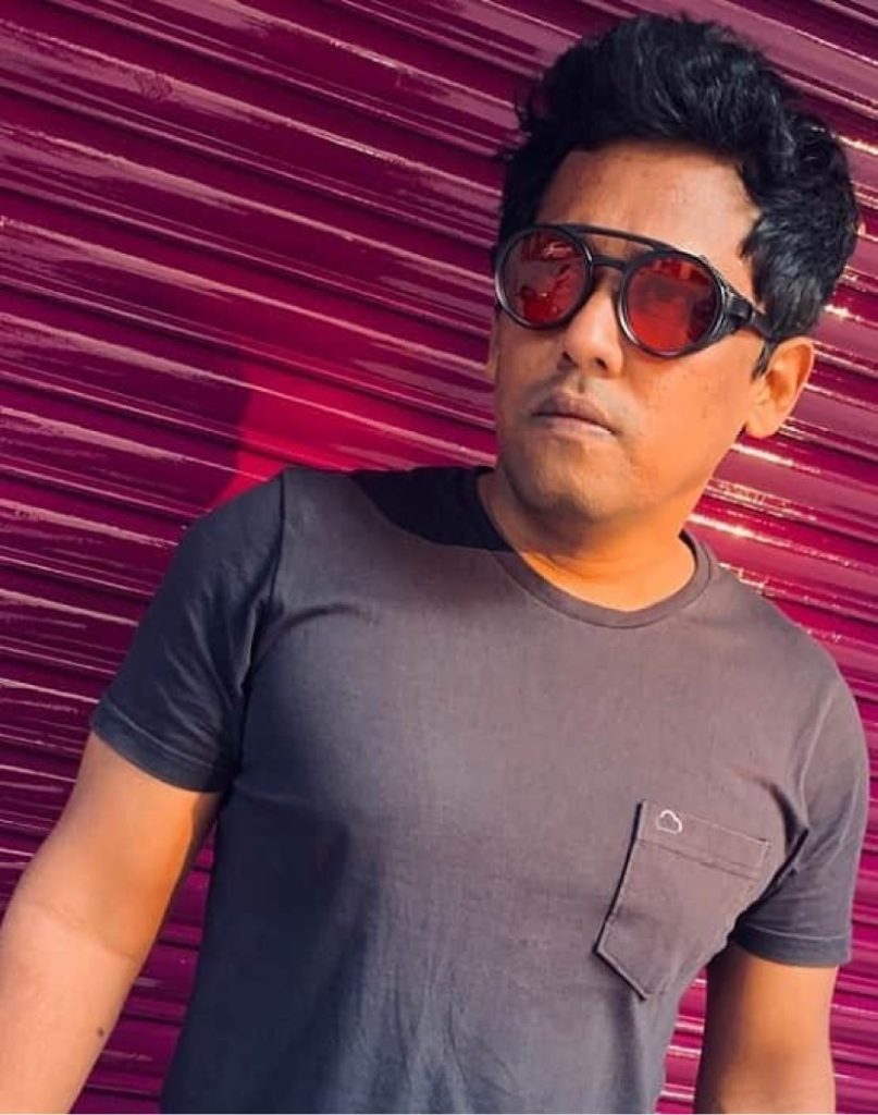 Indian DJ and producer Psy Inertia leaning against metal shutters and wearing sunglasses, colour image.