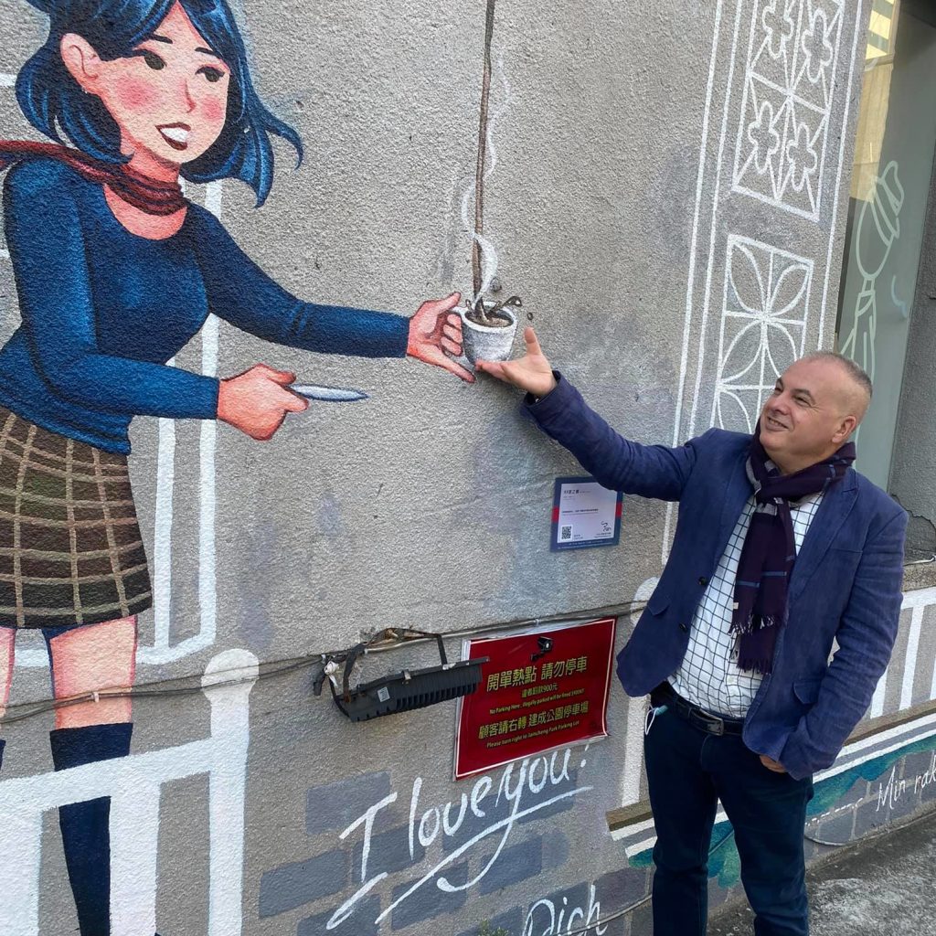 Italian Dj and producer Marco Mei taking a cup of coffee from Painting on wall with a female waiter, colour image.