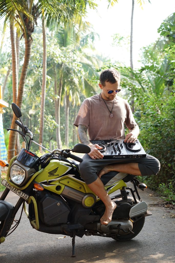 Belarusian DJ Igor Hush Sitting on Motorbike In South East Asian Jungle with a DJ controller on his lap.