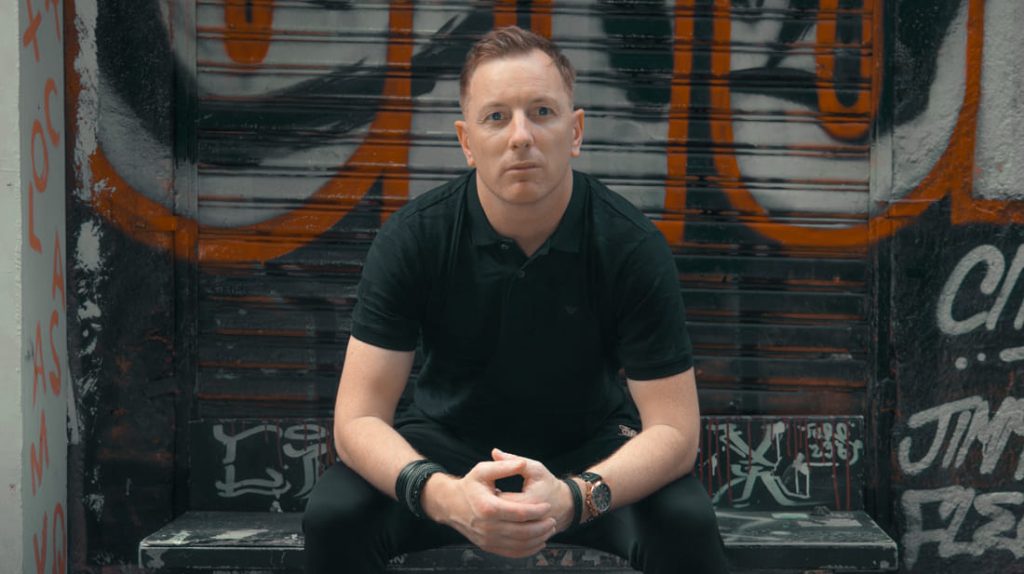 British DJ and producer Jon Connor sitting on bench looking at camera, colour image.
