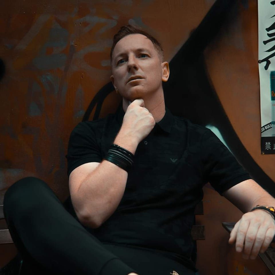 British DJ and producer Jon Connor sitting with hand on his neck, colour image.
