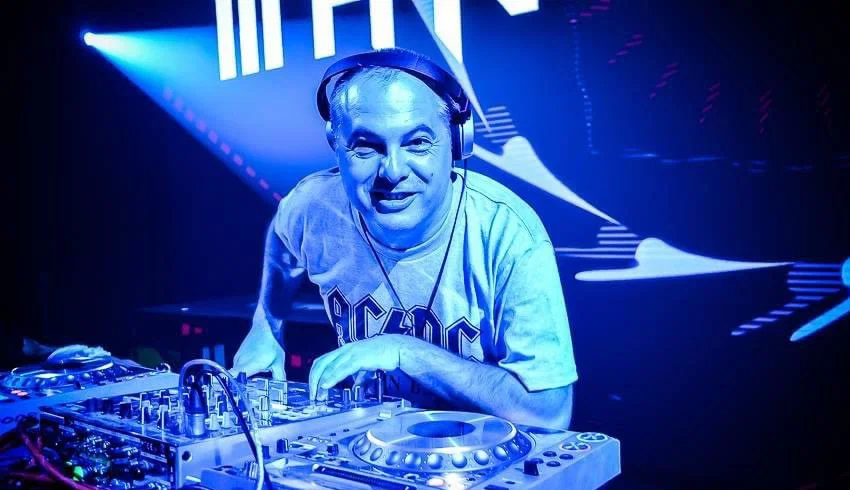 Marco Mei, Italian DJ and producer, smiling at the camera behind Pioneer CDJs wearing an AC/DC t-shirt in a blue-lit club.