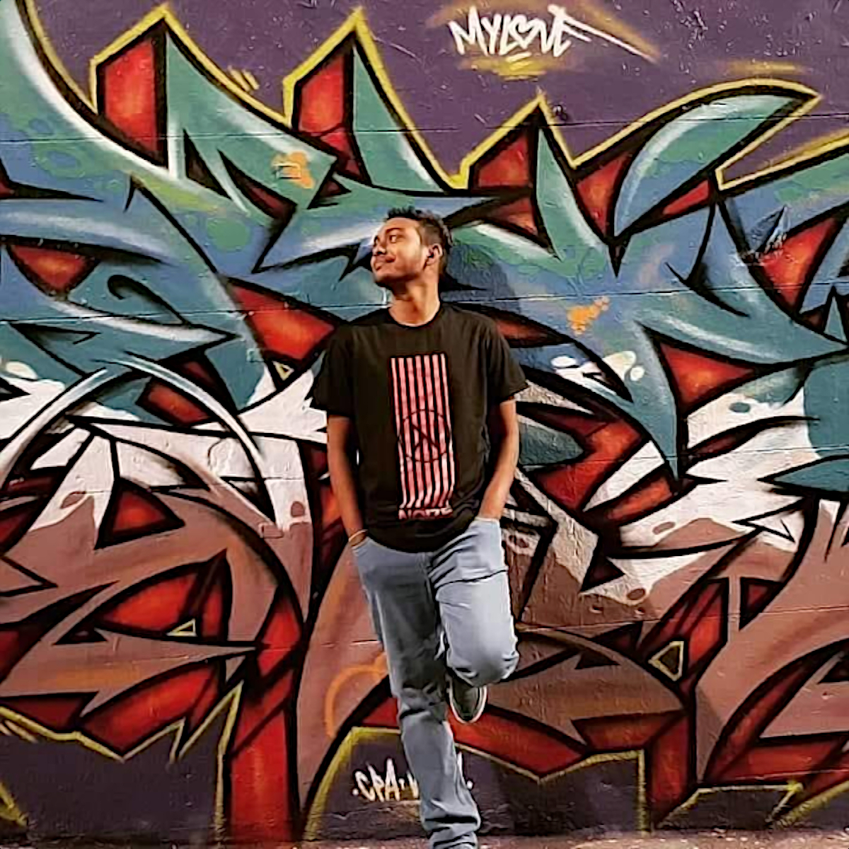 Sri L:ankan DJ and Producer KaZkO standing in front of graffiti wall with hands in pockets, colour image.
