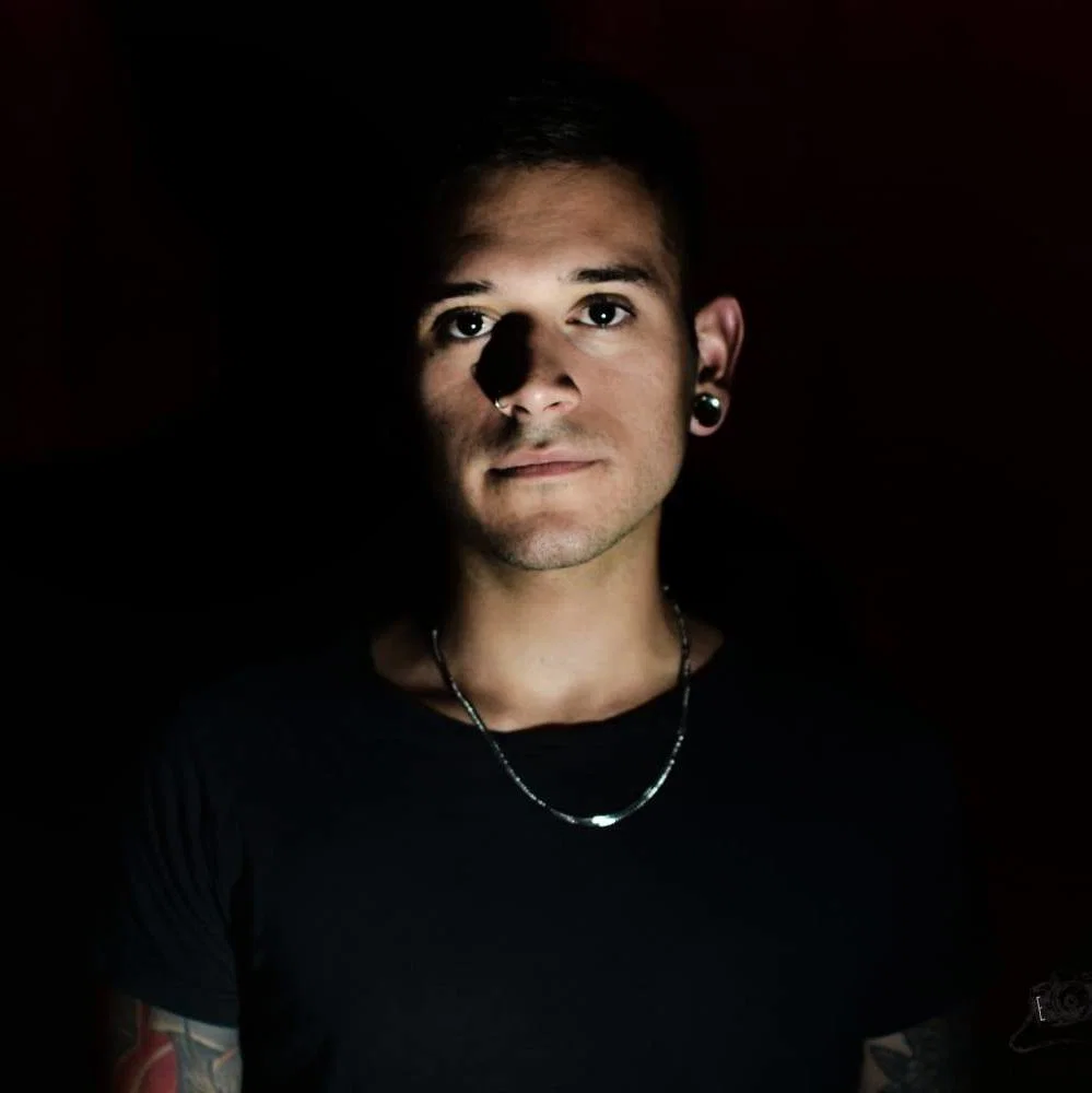 Portrait of Matías Ocaño wearing a black T-shirt and chain necklace against a dark background, with dramatic lighting across his face and visible tattoos on his arms.