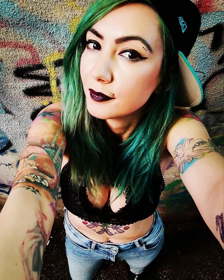Romanian DJ an producer Deysa standing with back to wall taking a selfie with green hair and baseball cap facing rear.
