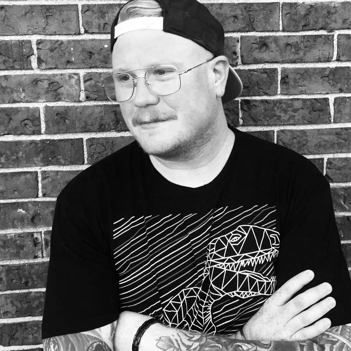 Guest DJ Coach in a black and white portrait, wearing glasses and a backwards cap, standing with arms crossed against a brick wall backdrop.