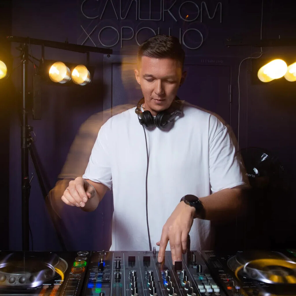 AKIN K performing on DJ equipment in a white T-shirt with headphones around his neck, under warm stage lights.