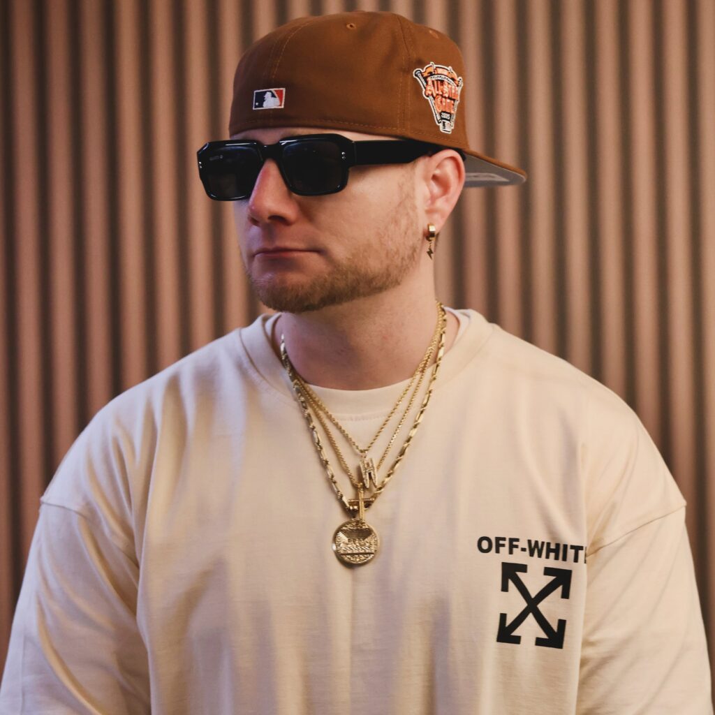 Hatch Wiseguy wearing black sunglasses, a brown baseball cap, a white T-shirt and layered gold chains against a softly lit indoor background.