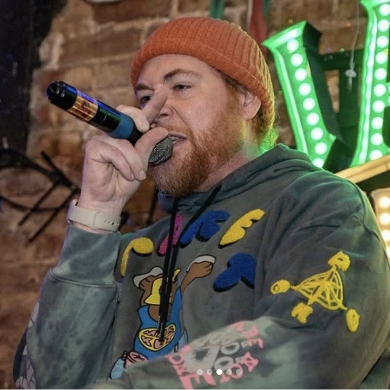 John Brown The Rapper wearing an orange beanie and colorful hoodie performs with a microphone in front of a neon sign.