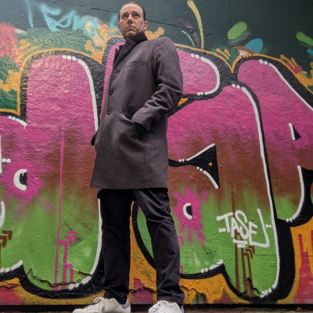 Blaze DJ in a dark coat and white sneakers stands confidently in front of vibrant graffiti art.