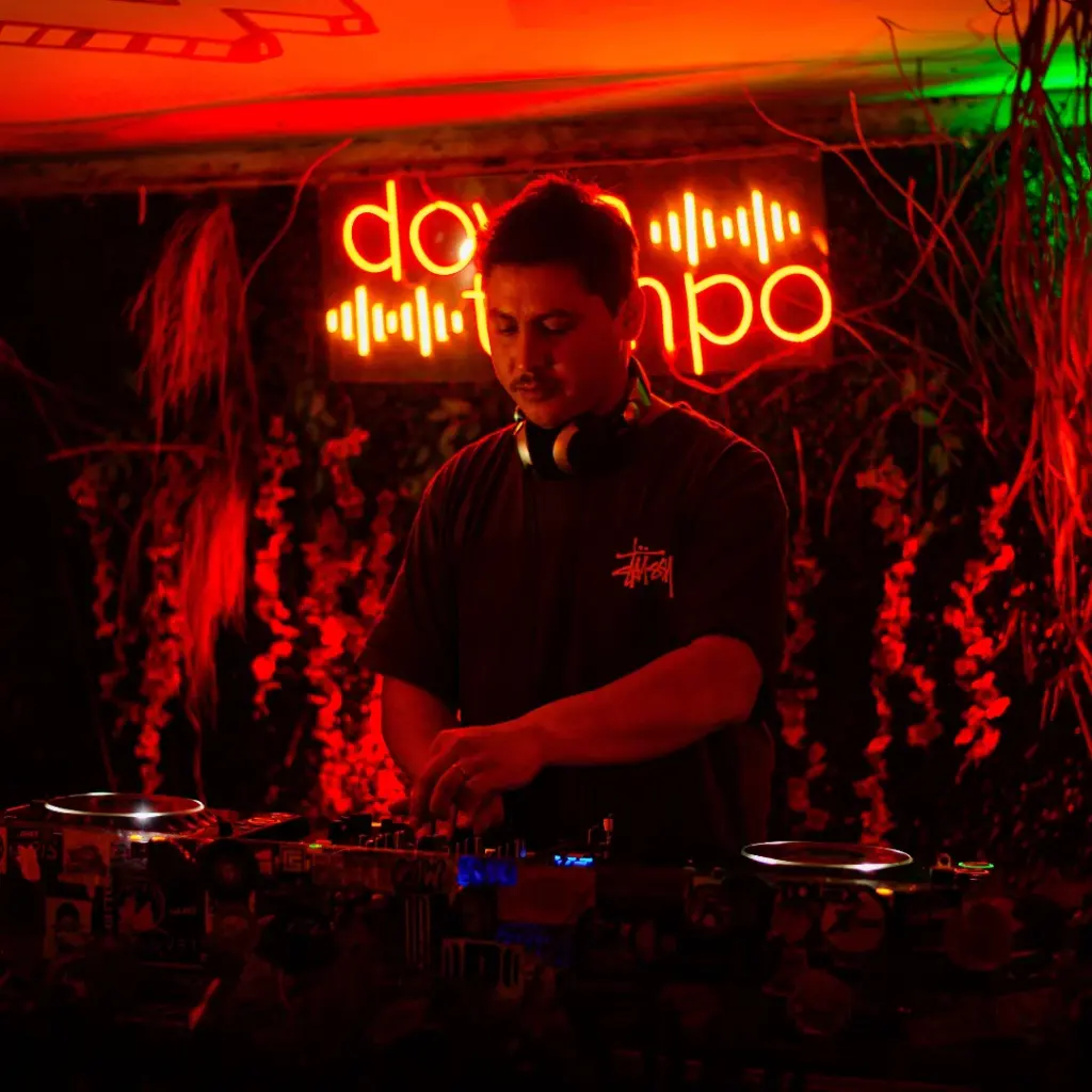 Argentinian Progressive House and Organic House DJ and producer Franco Mohr performing behind DJ decks, wearing a black T-shirt with headphones around his neck under vibrant red lighting and a glowing neon sign.