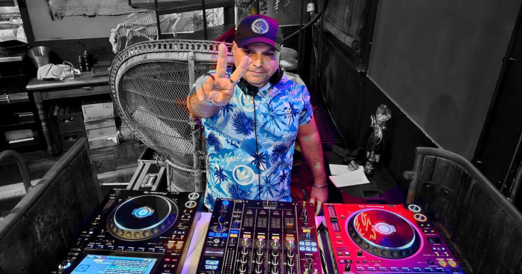Italian DJ and producer Carmelo Carone stands behind Pioneer DJ decks and a mixer, wearing a colourful blue tropical-print shirt and black cap while making a peace sign towards the camera. The photograph combines a colour portrait with a black-and-white background.