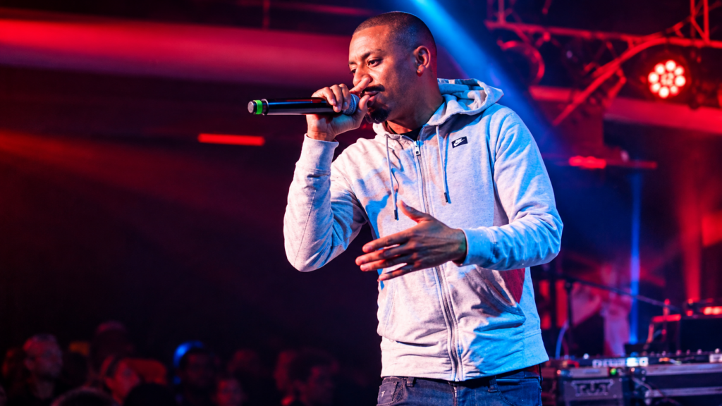 DJ King Flow guest American underground Hip Hop MC Curtis Coke from Rochester, New York, performing live with a microphone, wearing a grey Nike hoodie under vibrant red and blue stage lighting.
