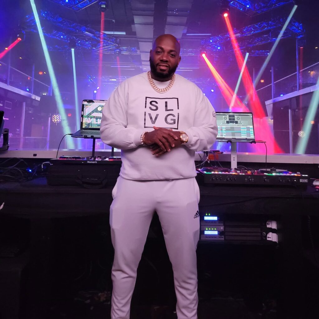 DJ Chuck T stands in front of DJ equipment at a nightclub, wearing a white tracksuit and gold chain, with colourful red, blue and purple stage lighting behind him.