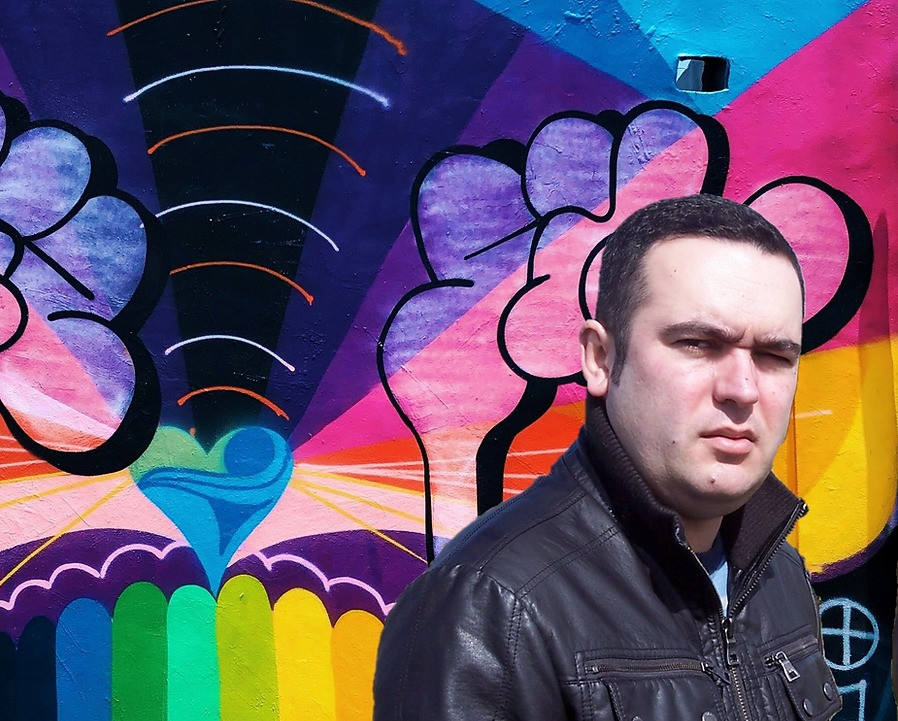 DJ Joshua wearing a dark jacket in front of a colourful graffiti wall.