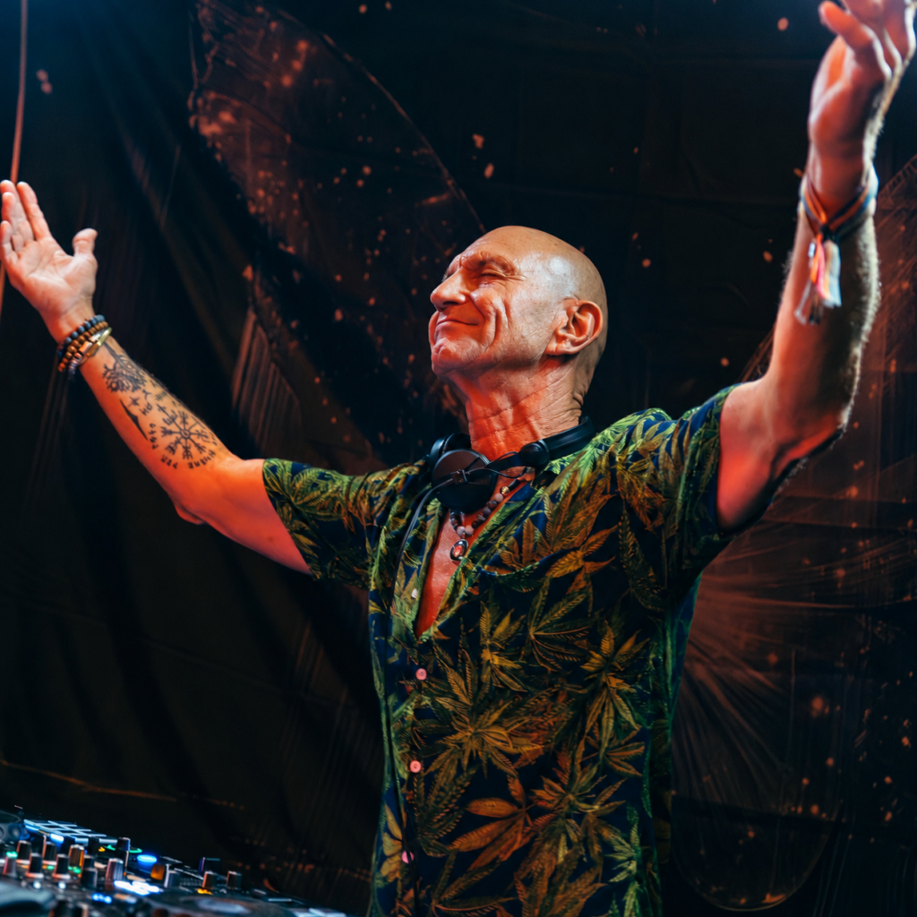 Belgian DJ Massimo Conecto, also known as Dirk Stommels, performing behind DJ decks with his arms raised, wearing a green tropical-print shirt and headphones around his neck under warm stage lighting.